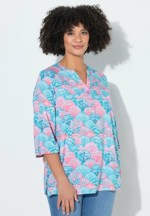 Kobieta o kręconych włosach, ubrana w kolorową bluzkę z motywem muszelek w kolorze różowym, niebieskim i morskim z rękawami do połowy przedramienia oraz niebieskie dżinsy.