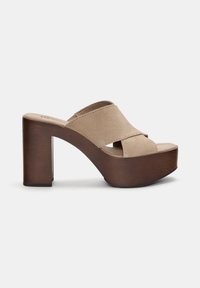 Beige ruskind højhælede mules med en platform og en brun træblokhæl. Har krydset stropper og en glat tekstureret overflade.