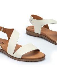Sandalias de cuero blancas con dos correas anchas, una correa trasera en el tobillo y una suela marrón. Textura suave con acentos mínimos y perfil plano.