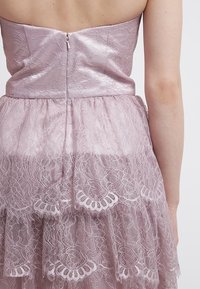 Spetsklänning i ljusrosa med en åtsittande satinöverdel och ett lager av spetskjol. Har en bakre dragkedja och detaljer med delikat spetskant.