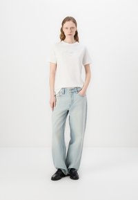 Cămașă din bumbac bej deschis cu un design de text iridescent. Purtați cu blugi largi de culoare albastră deschisă și pantofi din piele negri. Fundal minimalist.
