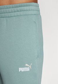 Šviesiai mėlynos joggingo kelnės, pagamintos iš minkštos medžiagos, turi raukšlėtą juosmenį ir išsiuvinėtą baltą Puma logotipą ant kairiojo šlaunies.