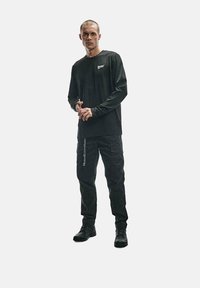 Czarna koszula z długim rękawem z logo, w połączeniu z czarnymi spodniami cargo z bocznymi kieszeniami i oznakowaniem. Model nosi czarne buty.