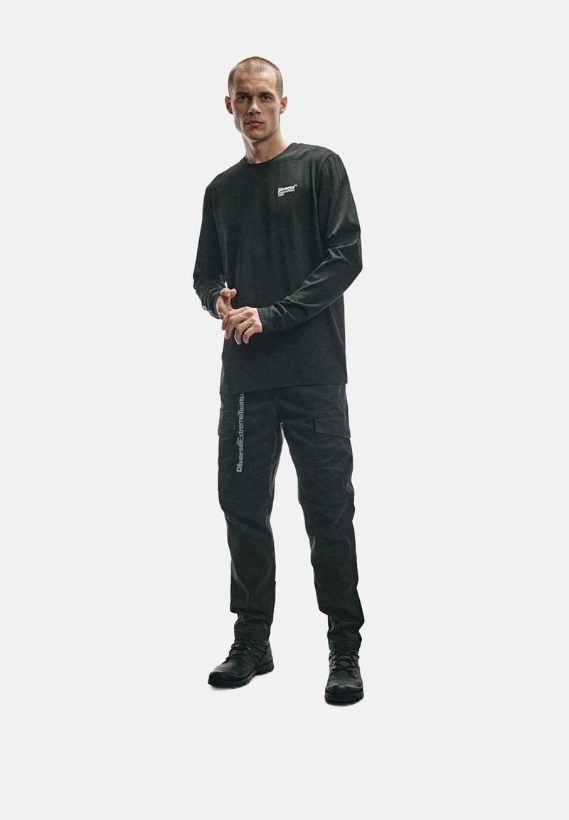 Czarna koszula z długim rękawem z logo, w połączeniu z czarnymi spodniami cargo z bocznymi kieszeniami i oznakowaniem. Model nosi czarne buty.