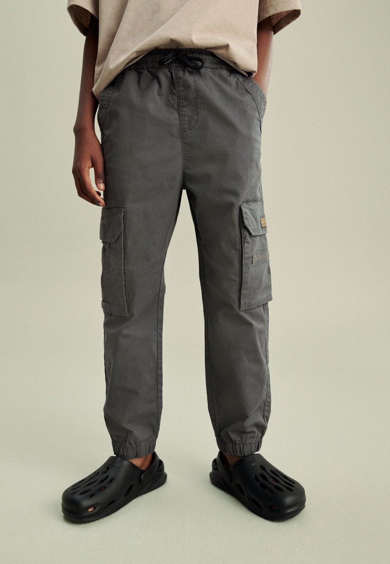 Personne portant un pantalon cargo gris foncé avec des poignets élastiques et des chaussures slip-on noires avec des trous de ventilation.