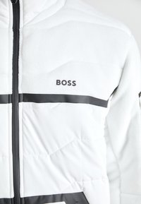 Bílá fleece bunda s černými akcenty a zipovým uzávěrem, s prošívanou texturou a názvem značky "BOSS" na hrudi.