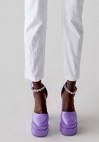 Scarpe a piattaforma viola con una finitura liscia, dotate di una cintura nera e un braccialetto decorativo con gemme trasparenti. Indossate con pantaloni bianchi a fantasia.