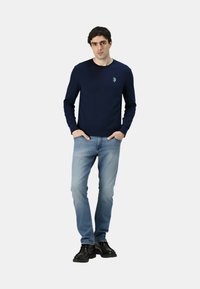 Felpa a maniche lunghe blu navy con girocollo e piccolo logo, abbinata a jeans azzurri e scarpe nere. Design pulito e casual.
