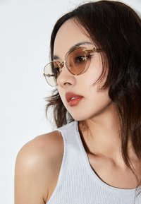 Occhiali da sole a forma di occhio di gatto con accenti dorati e lenti marroni. Presentano una sottile montatura in metallo e un design angolato, indossati da un modello con capelli mossi.