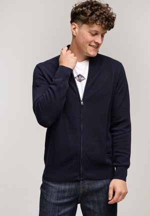 Granatowy kardigan na zamek błyskawiczny wykonany z teksturowanej tkaniny, z długimi rękawami i prostym wzorem. Nosi się go na białej koszulce, w połączeniu z ciemnymi dżinsami.