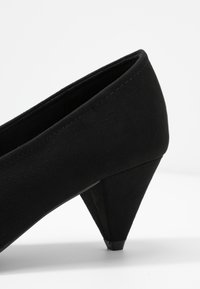 Chaussure noire à talons hauts avec une tige douce de type daim, un talon triangulaire incliné et une texture lisse avec des détails de couture subtils.