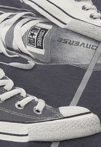 Grafik av grå Converse-sneakers med vita snören och sulor. Har texturerat tyg och mönsterdetaljer på en mörk bakgrund.