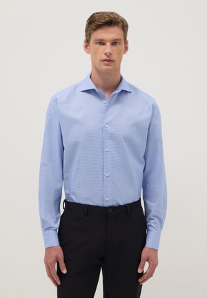 Joven con camisa azul clara de botones y mangas largas, y pantalones negros, de pie frente a un fondo claro y liso.