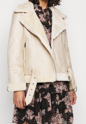 Giacca di shearling beige con grande colletto, texture trapuntata, tasche con zip e vita cinturata. Indossata sopra un vestito floreale.