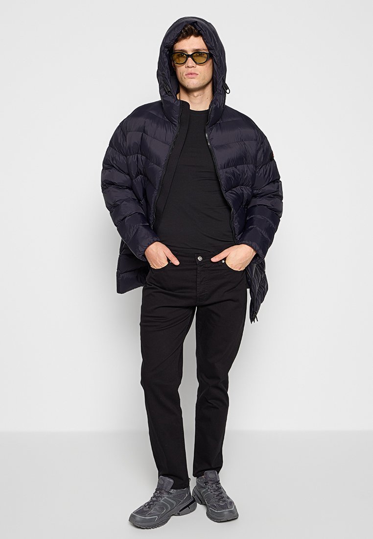 Gevoerde marineblauwe jas met capuchon, zwart t-shirt, zwarte broek en grijze sneakers. Strakke uitstraling en zachte textuur, met een casual pasvorm.