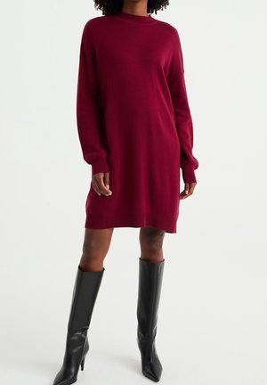 Femme portant une robe pull bordeaux à manches longues, longueur genou, associée à des bottes noires à talons montant jusqu'aux genoux, debout devant un fond uni.