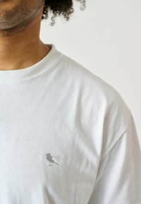 Weißes Baumwoll-T-Shirt mit Rundhalsausschnitt. Auf der linken Brust befindet sich ein kleines graues Vogel-Logo. Der Stoff wirkt weich mit einer leichten Textur.