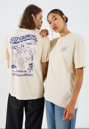 Camisetas de algodón beige con estampados gráficos; una presenta la frase "SIGUE CRECIENDO" y diseños florales, la otra tiene un pequeño logotipo de sol en el pecho.