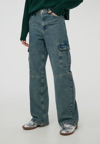 Luźne dżinsy cargo w jasnoniebieskim deniźie o wysokim stanie, z dwoma kieszeniami po bokach i prostym krojem nogawki. W połączeniu ze srebrnymi butami.