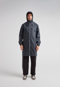 Mörkgrå vattentät regnjacka med huva, framsida med dragkedja och elastiska muddar, bärs över svarta byxor och vita sneakers. Slät textur.