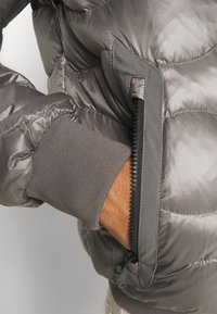 Giacca imbottita grigia con finitura liscia e lucida, polsini elastici e una tasca con zip sul lato. Tessuto testurizzato visibile.