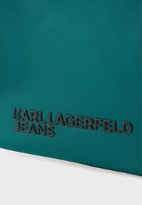 Modrá nylonová taška na plece s vyrazeným čiernym textom "KARL LAGERFELD JEANS," hladkou textúrou a elegantným dizajnom. Žiadne viditeľné kovania ani doplnky.