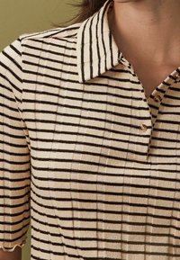 Kremowa koszulka polo z wąskimi brązowymi paskami, żebrowaną teksturą i kołnierzykiem z trzema guzikami. Krótkie rękawy.