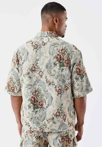 Camicia a maniche corte in tessuto testurizzato presenta un motivo floreale con tonalità di rosso, verde e blu su uno sfondo chiaro.