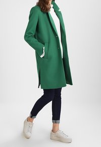 Eine Frau in einem leuchtend grünen Mantel steht selbstbewusst da, trägt ein weißes Oberteil, dunkle Jeans und weiße Sneaker, mit den Händen in den Taschen.