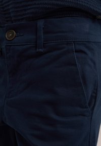 Tamsiai mėlynos kelnės, pagamintos iš tekstūrinio medvilnės audinio, su priekiniais kišenėmis, vario mygtuku ir pritaikytu siluetu su minimaliais raukšlėmis.