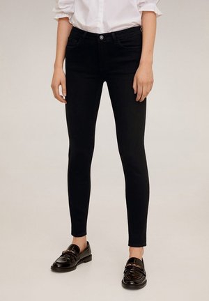 Mujer vistiendo jeans pitillo negros con una camisa blanca y mocasines negros contra un fondo neutro.