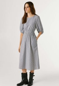 Žena u midi haljini sivo-bijelih pruga s puf rukavima i crnim čizmama do sredine lista, stoji s jednom rukom u džepu.