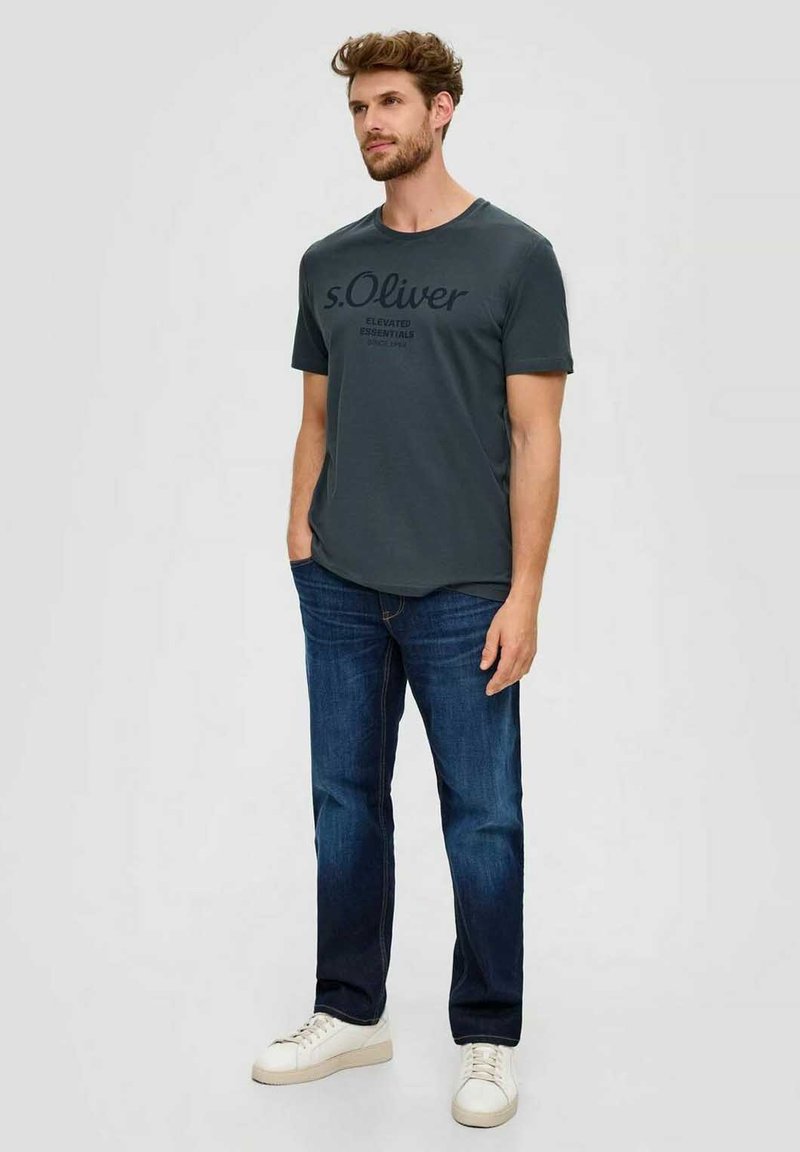 Dark gray t-shirt with a crew neck and short sleeves, featuring black printed text. Paired with dark blue jeans and white sneakers.