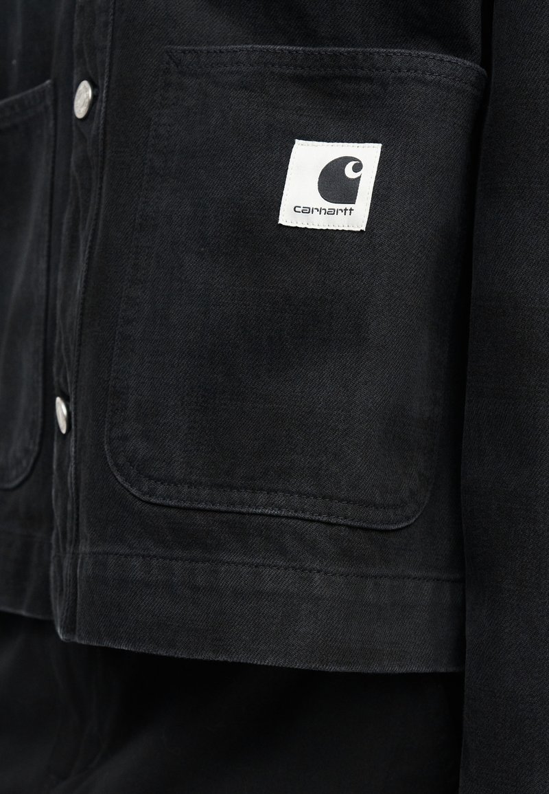 Black Carhartt jacket pocket with white square logo patch and metal snap buttons on left side.