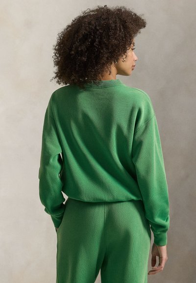 Γυναίκα με σγουρά μαλλιά στέκεται πλαγίως φορώντας πράσινο μακρυμάνικο τοπ και ταιριαστό πράσινο παντελόνι μπροστά σε ένα απλό φόντο.