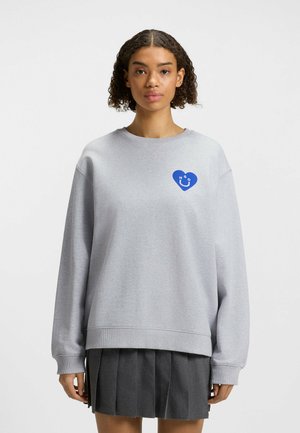 Gray sweatshirt featuring a blue heart-shaped logo on the left chest. Loose fit with long sleeves and ribbed cuffs. Soft, textured fabric.