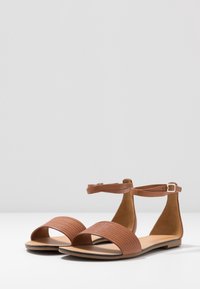 Ruske platte sandaler med teksturert bred rem og tynn ankelrem. Åpen tå-design med en glatt såle. Minimalistisk stil og sikker spenne.