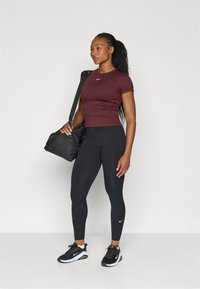 Top atlético ajustado de color burdeos, leggings negros y zapatillas negras. El modelo sostiene un bolso de gimnasio negro, que presenta un logo blanco en la parte superior.