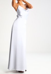 Kvinna i en vit ärmlös satängklänning med en draperad halsringning, stående med en arm lyft mot en enfärgad ljus bakgrund.