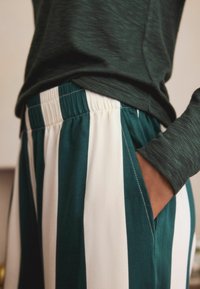Grün gestreifte Stoffhosen mit einem breiten elastischen Bund, Seitentaschen und einer glatten Textur, ergänzt durch ein dunkelgrünes Langarmoberteil.
