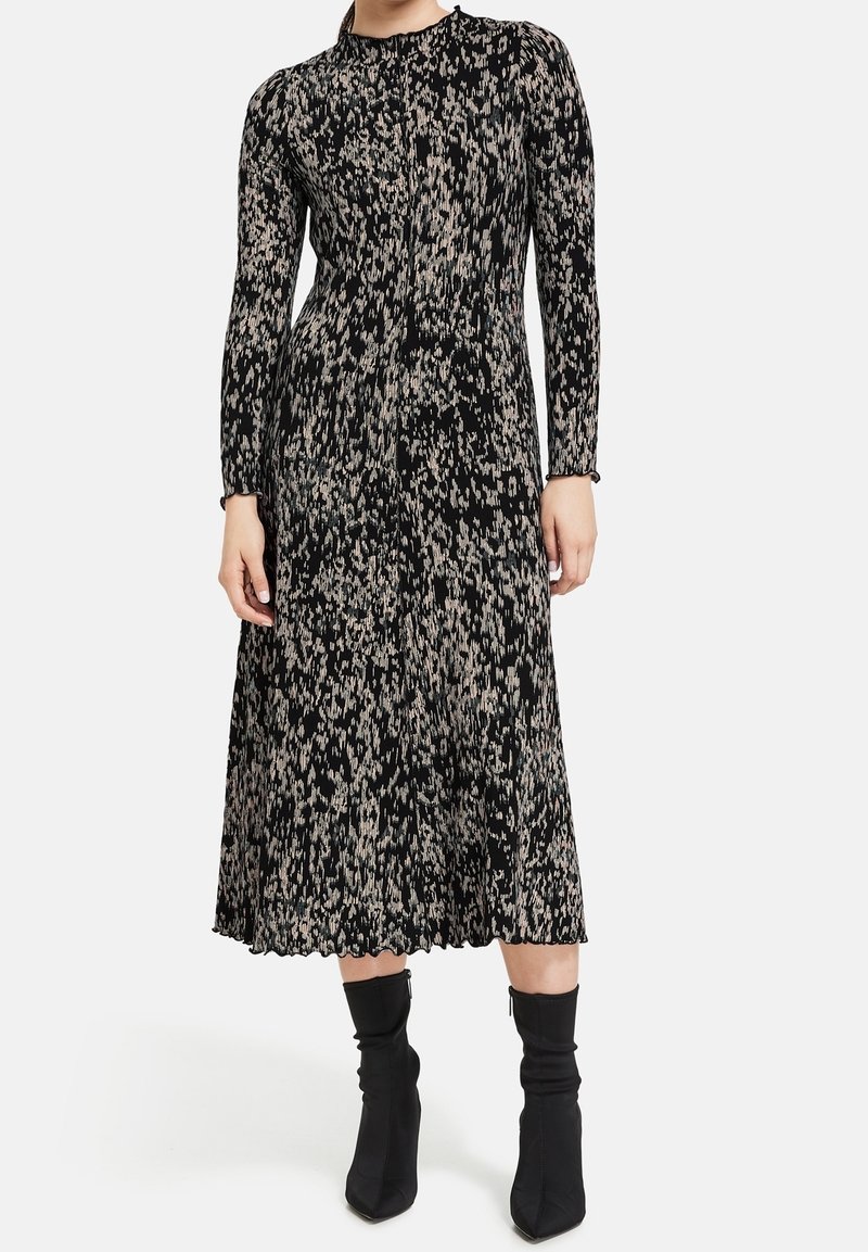 Femme portant une robe midi noire à manches longues avec un imprimé abstrait beige et des bottines chaussettes noires à bout pointu, debout sur un fond blanc.