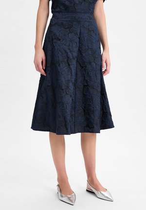 Femme portant une jupe midi bleu marine à texture florale et des ballerines argentées à bride arrière à bouts pointus, debout sur un fond blanc.