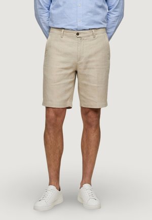 Homme portant un short beige mi-cuisses, une chemise bleu clair à boutons et des baskets blanches, debout avec les mains dans le dos.