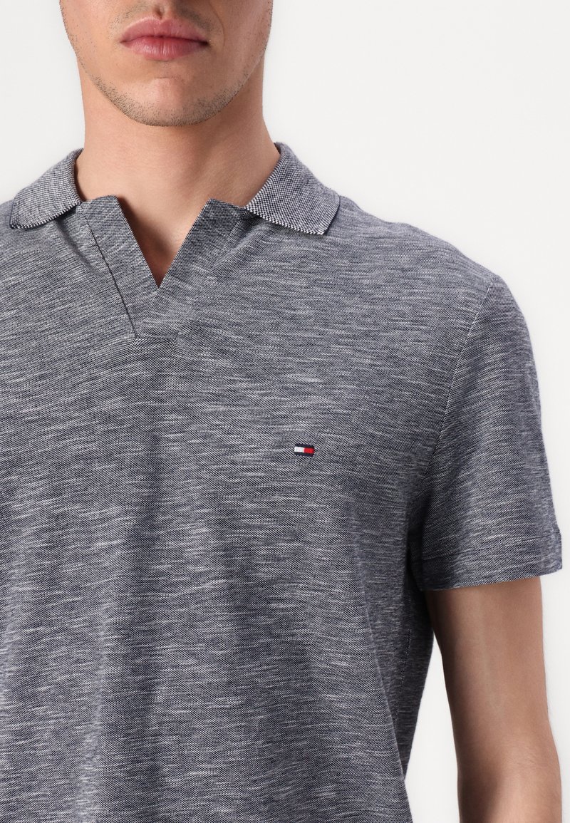 Polo gris con una tela texturizada, con cuello en V y mangas cortas. Pequeño logo en rojo, blanco y azul en el lado izquierdo del pecho.