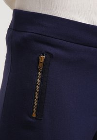 Marine-blauwe stoffen broek met een goudkleurige ritszakdetail. Glad materiaal met een getailleerde pasvorm bij de taille.