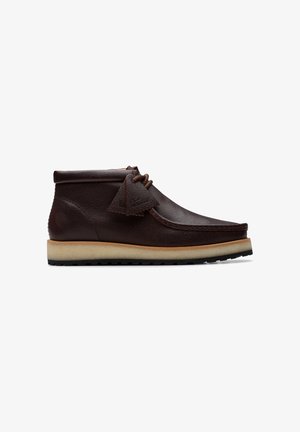 Stivaletto in pelle marrone scuro con suola in crepe, cucitura a moc toe, chiusura con lacci e etichette in pelle per i lacci su sfondo bianco.