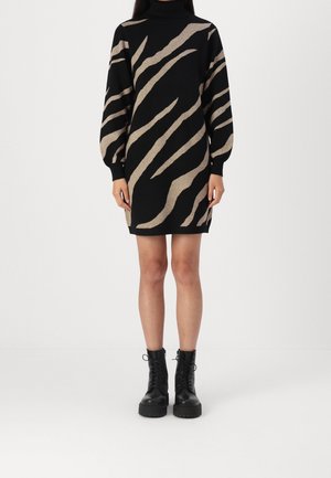 Frau trägt ein langärmliges Rollkragenkleid mit schwarzen und beigen Zebramustern und schwarze, schnürbare Knöchelstiefel, steht vor einem weißen Hintergrund.