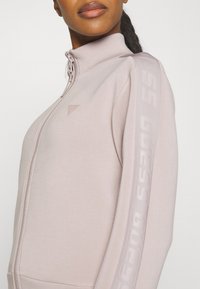 Light pink zip-up sweatshirt with high collar, featuring tonal "GUESS" logo along the sleeves, made from smooth, soft fabric.