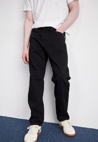 Osoba v černých kalhotách a bílých teniskách stojí proti bílé zdi, s jednou rukou v kapse na modré koberec.