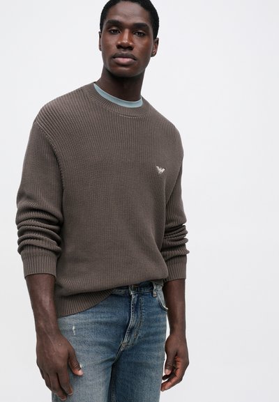 Rjava pletena pulover s rebrastim vzorcem, okroglo izrezano ovratnikom in manšetami. Vključuje majhen logotip na prsih, oblečeno z zbledelimi modrimi kavbojkami.